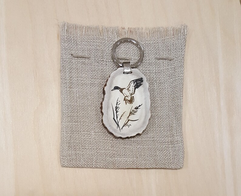 May include: A brown antler keychain with a painted image of a duck in flight. The antler is oval shaped and has a silver key ring attached.