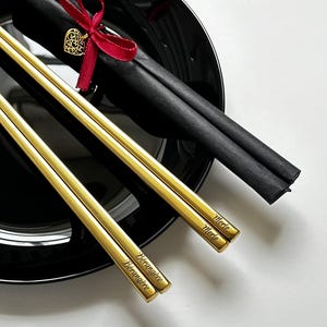 Puede incluir: Palillos dorados con nombres grabados, colocados sobre un plato negro. Los palillos se acompa&ntilde;an de una funda de papel negro, atada con una cinta roja y un dije de coraz&oacute;n dorado. Los nombres "Merle" y "Berangere" est&aacute;n grabados.