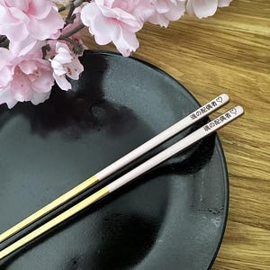 Puede incluir: Un par de palillos con puntas doradas y cuerpos rosa claro, sobre un plato negro. Los palillos tienen texto japon&eacute;s y un s&iacute;mbolo de coraz&oacute;n. Flores rosas en el fondo.