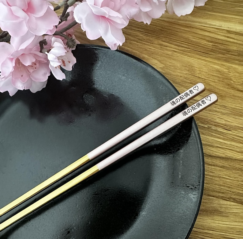 Puede incluir: Un par de palillos con puntas doradas y cuerpos rosa claro, sobre un plato negro. Los palillos tienen texto japon&eacute;s y un s&iacute;mbolo de coraz&oacute;n. Flores rosas en el fondo.