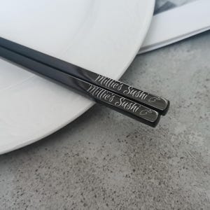 Puede incluir: Un par de palillos chinos negros con el texto "Millie's Sushi" grabado en cursiva. Los palillos est&aacute;n sobre un plato blanco, con un fondo gris.