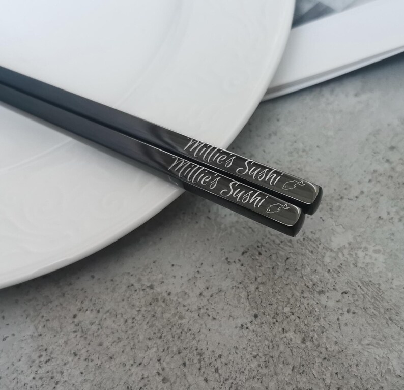 Puede incluir: Un par de palillos chinos negros con el texto "Millie's Sushi" grabado en cursiva. Los palillos est&aacute;n sobre un plato blanco, con un fondo gris.