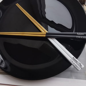 May include: A pair of chopsticks with black and gold handles. The chopsticks are engraved with the names "Aalana Phu" and the date "31.12.2022".