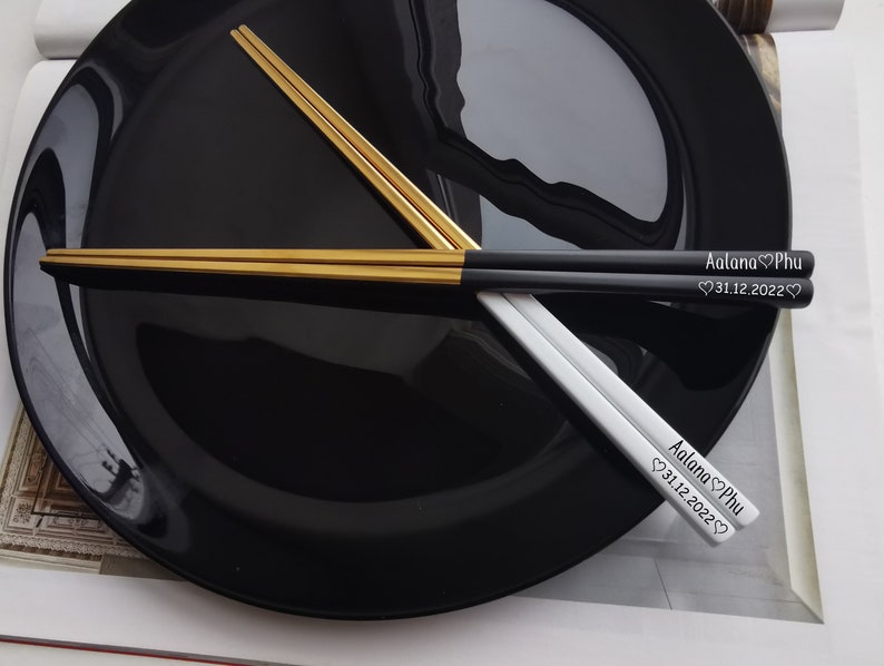 May include: A pair of chopsticks with black and gold handles. The chopsticks are engraved with the names "Aalana Phu" and the date "31.12.2022".