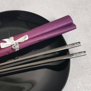 May include: Two silver chopsticks with the names "Tarlea" and "Jorge" engraved on them, resting on a black plate with a purple and white napkin tied with a ribbon and a heart charm.