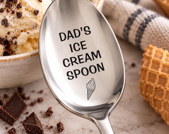 May include: A polished stainless steel spoon featuring the black engraved text DAD'S ICE CREAM SPOON and a small graphic of an ice cream cone in a waffle cone. The spoon has a smooth, reflective surface and a classic rounded bowl shape.
