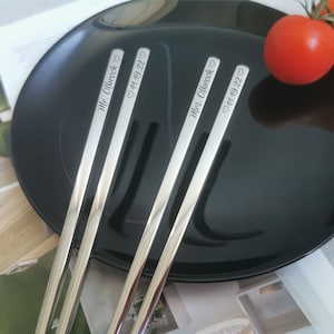 Puede incluir: Cuatro palillos plateados descansan sobre un plato negro, con un tomate rojo en el fondo. Cada palillo est&aacute; grabado con un nombre y una fecha: "Mr. Olbrook 11.19.22" y "Mrs. Olbrook 11.19.22".