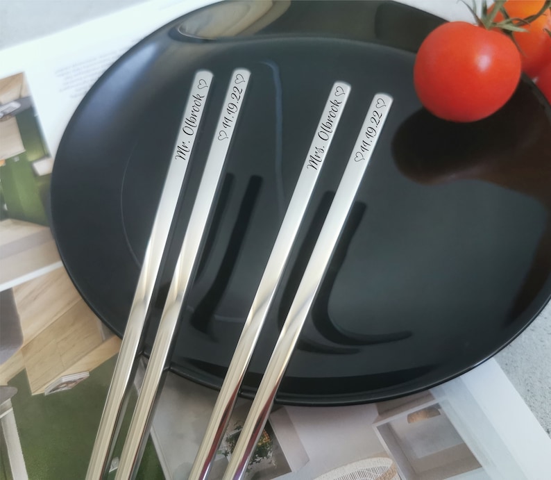 Puede incluir: Cuatro palillos plateados descansan sobre un plato negro, con un tomate rojo en el fondo. Cada palillo est&aacute; grabado con un nombre y una fecha: "Mr. Olbrook 11.19.22" y "Mrs. Olbrook 11.19.22".