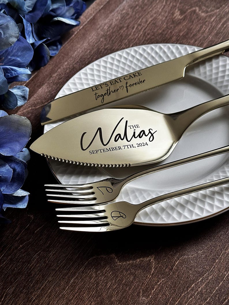 May include: A gold-colored wedding cake knife and server set with the words "Let's eat cake together forever" and "The Walias September 7th, 2024" engraved. Two forks are also included. The set is on a white plate.