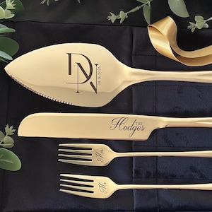 May include: A gold-toned cake serving set consisting of a serrated cake server, a knife, and two forks. The server is engraved with the initials DN and the date 08.16.2026. The knife features the text The Hodges, while the forks are engraved with Mrs. and Mr.