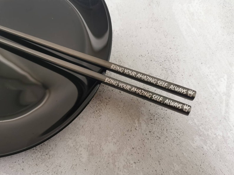 May include: A pair of black chopsticks with the words "Being your amazing self, always" engraved on them. A small crown symbol is also engraved on each chopstick.