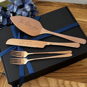 May include: A copper-colored cake serving set with a cake server, knife, and two forks, presented in a black box with a blue ribbon. The cake server is engraved with "The Ribnickys" and a date. The knife reads "Let's eat cake together-forever."