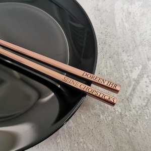 Puede incluir: Palillos chinos de oro rosa con texto grabado, sobre un plato negro. Los palillos tienen grabadas las palabras "DOREEN IHR SEINE CHOPSTICKS". Un accesorio de mesa elegante y sofisticado.