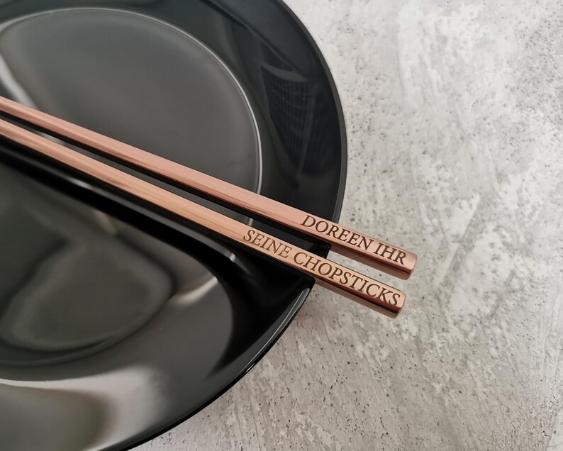 Puede incluir: Palillos chinos de oro rosa con texto grabado, sobre un plato negro. Los palillos tienen grabadas las palabras "DOREEN IHR SEINE CHOPSTICKS". Un accesorio de mesa elegante y sofisticado.
