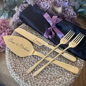 May include: A gold-colored wedding cake server set with a cake server, knife, and two forks. The cake server is engraved with "Chase & Montana", the knife with "Lorenz", and the forks with "Mr". The set is on a woven mat with a black cloth and purple ribbon.