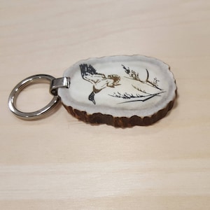 May include: A key chain made from a piece of antler with a burned image of a bird in flight. The antler is light brown and the bird is black. The key chain has a silver ring.