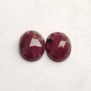 May include: Two oval-shaped, faceted, deep red gemstones. The gems have a polished surface with multiple facets that catch the light. The gemstones are set against a white background.