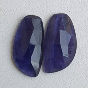 Peut inclure: Deux pierres précieuses en forme de goutte d'eau, facettées, d'un bleu indigo profond. Les gemmes ont une surface polie et réfléchissante, avec de multiples facettes captant la lumière. Les pierres sont posées sur un fond blanc.