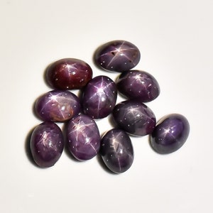 May include: A collection of oval-shaped, star-effect gemstones in varying shades of purple and burgundy. Each stone displays a six-rayed star pattern, with a polished, reflective surface. The gemstones are arranged on a white background.