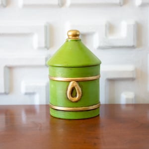 Op de afbeelding: Een groene decoratieve pot met gouden accenten. De pot heeft een cilindrisch lichaam, een deksel en een gouden lusdetail. De pot is ongeveer 15 cm hoog.