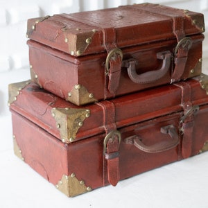 May include: Two stacked, vintage-style, rectangular suitcases. The suitcases are a deep reddish-brown with brass-colored corner protectors and buckles. Each suitcase has a handle and leather straps.