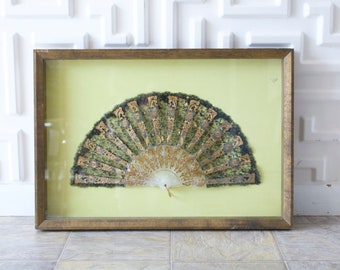 May include: Framed vintage fan with a gold-toned wooden frame. The fan features a detailed design with gold and green accents on a light green background. The fan is displayed in a glass-covered frame, adding a touch of elegance.