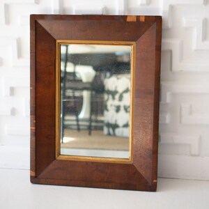 May include: A rectangular wooden framed mirror with a gold inner frame. The dark brown frame has a geometric design. The mirror reflects a room with white walls and a patterned curtain.