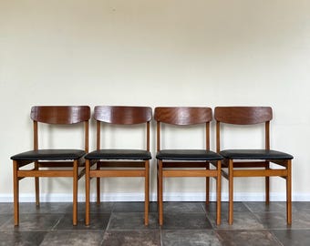 May include: Four mid-century modern chairs. Each chair has a curved, dark wood backrest, a black seat, and a light brown wooden frame. The chairs are arranged in a row against a neutral-colored wall and sit on a tiled floor.