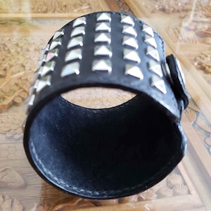 May include: A black leather cuff bracelet with silver pyramid-shaped studs. The bracelet is adjustable and has a buckle closure.
