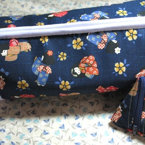 May include: A navy blue cylindrical bag and a triangular coin purse. Both items feature a repeating pattern of geishas in colorful kimonos, yellow flowers, and white blossoms. The bag has a white trim and a lavender handle.