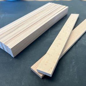 Op de afbeelding: Een stapel van 10 natuurlijke houten knutselstokjes, ongeveer 6 mm dik en 30 cm lang, en twee extra stokjes, een bovenop de andere, liggen op een donkergrijs oppervlak.