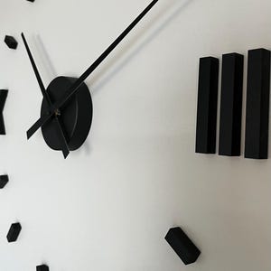 Könnte beinhalten: Eine moderne, übergroße Wanduhr mit schwarzen Zeigern und Stundenmarkierungen an einer weißen Wand. Die Uhr hat ein minimalistisches Design mit römischen Ziffern und eine elegante, moderne Ästhetik.