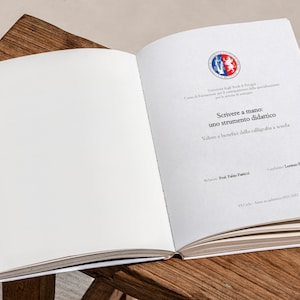 Può includere: Un libro aperto con una copertina bianca e una pagina di titolo che recita "Scrivere a mano: uno strumento didattico. Valore e benefici della calligrafia a scuola." Il libro è su una superficie di legno.