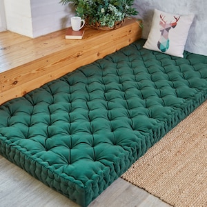 May include: A deep green tufted floor cushion with a button-tufted design. The cushion is placed on a wooden window seat and a natural fiber rug.