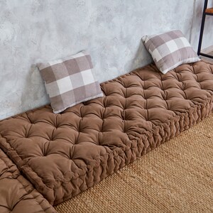 May include: Three brown floor cushions with tufted upholstery and a button-tufted design. The cushions are arranged in a row on a woven rug in front of a grey concrete wall. Two white and grey checkered throw pillows are on the cushions.