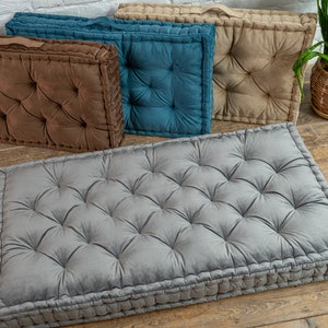 May include: Four floor cushions in different colours. The cushions are made of a soft, tufted fabric and have a rectangular shape. The cushions are arranged in a row on a wooden floor.