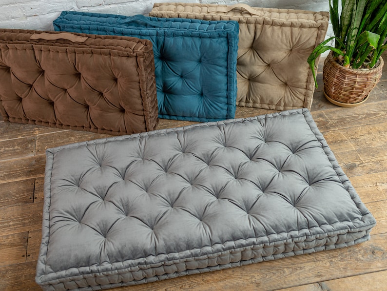 May include: Four floor cushions in different colours. The cushions are made of a soft, tufted fabric and have a rectangular shape. The cushions are arranged in a row on a wooden floor.