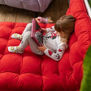 May include: A young girl sits on a red, tufted couch and uses a pink tablet. She is wearing a white long-sleeved shirt with a red and black Mickey Mouse design and grey pants.