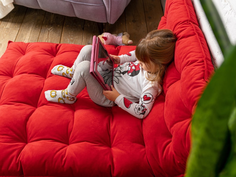 May include: A young girl sits on a red, tufted couch and uses a pink tablet. She is wearing a white long-sleeved shirt with a red and black Mickey Mouse design and grey pants.