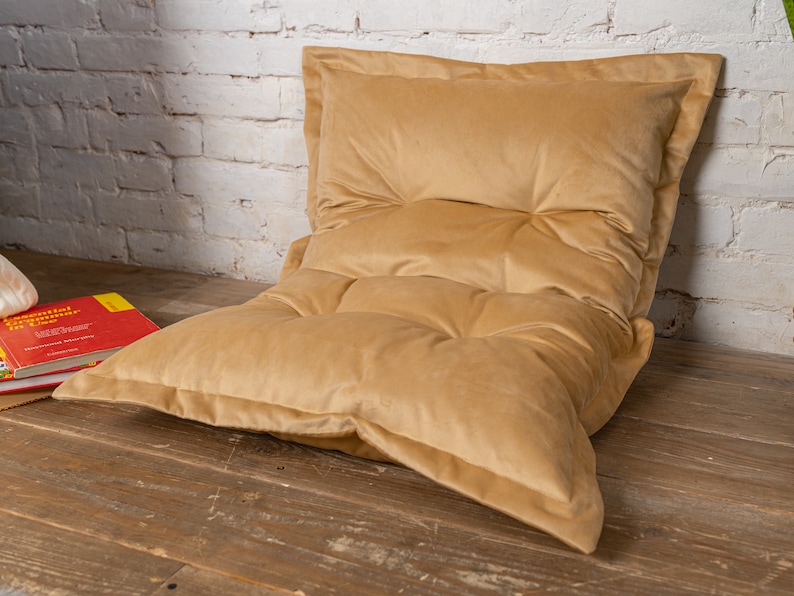 May include: A large, tan-colored, tufted floor cushion with a thick, plush fabric. The cushion is made of multiple layers of fabric, creating a comfortable and supportive seating option.