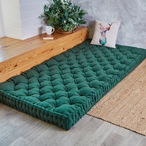 May include: A deep green, tufted floor cushion with a button-tufted design. The cushion is placed on a light wood floor and a natural fiber rug.