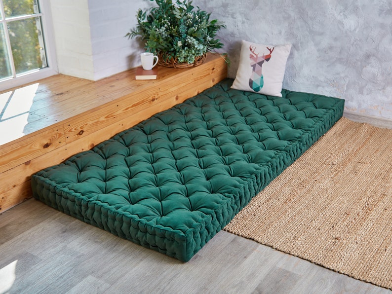 May include: A deep green, tufted floor cushion with a button-tufted design. The cushion is placed on a light wood floor and a natural fiber rug.