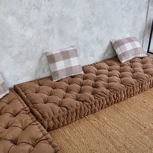 May include: Three brown floor cushions with tufted tops and white and gray checkered throw pillows. The cushions are arranged in a row on a woven rug in front of a gray concrete wall.
