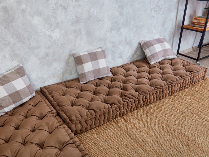 May include: Three brown floor cushions with tufted tops and white and gray checkered throw pillows. The cushions are arranged in a row on a woven rug in front of a gray concrete wall.