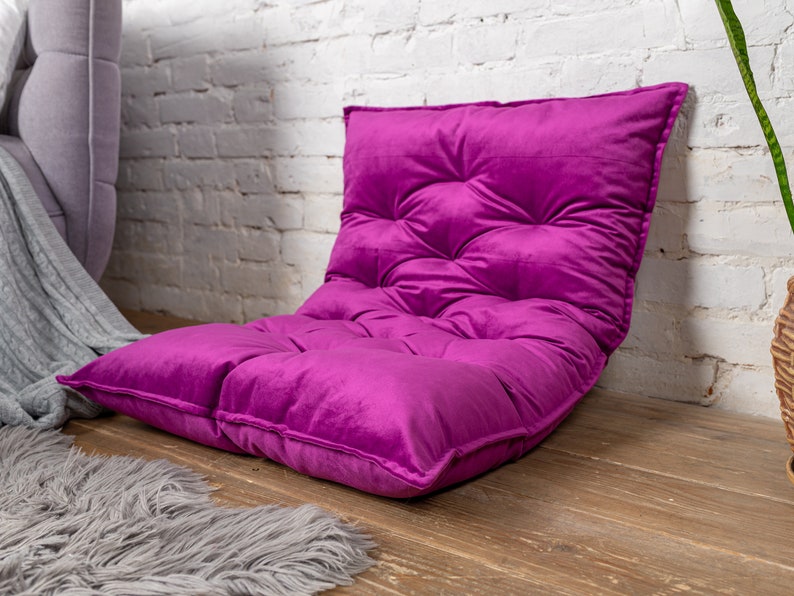 May include: A bright magenta floor cushion with a tufted design. The cushion is folded in half and is sitting on a wooden floor.