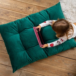 May include: A young person lies on a green velvet floor cushion and uses a tablet computer. The cushion has a tufted design.