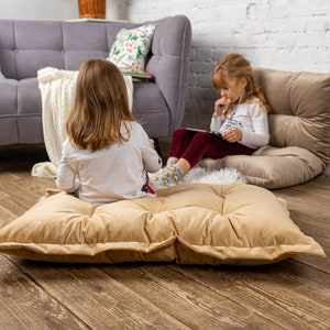 May include: Two children sitting on floor cushions in a living room. One child is sitting on a beige floor cushion and the other is sitting on a brown floor cushion. Both children are looking at a tablet.