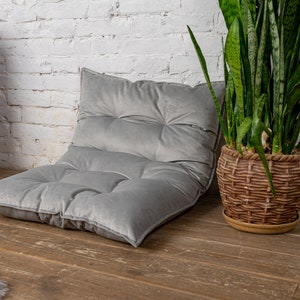 May include: A gray, tufted floor cushion with a thick, plush fabric. The cushion is sitting on a wooden floor in front of a white brick wall. A potted plant with long, green leaves is in the background.