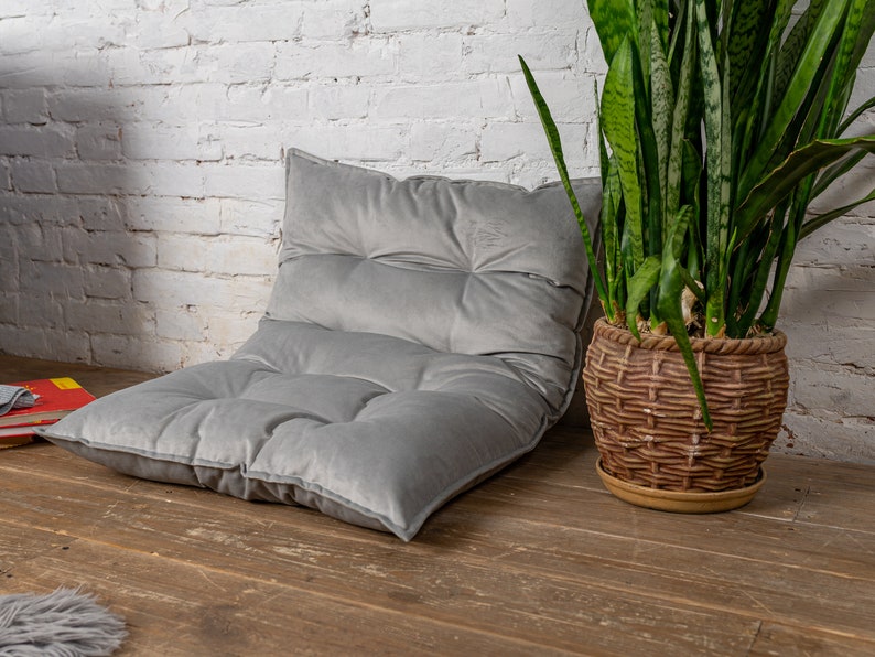 May include: A gray, tufted floor cushion with a thick, plush fabric. The cushion is sitting on a wooden floor in front of a white brick wall. A potted plant with long, green leaves is in the background.