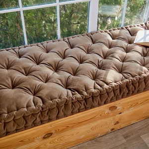 May include: A brown tufted cushion with a book on top, placed on a wooden window seat.